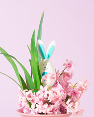 Egg shape bunny peeks out from the pink hyacinth flowers in the vase. Copy space. Pastel pink background. Frontal view. Minimal springtime holiday concept.
