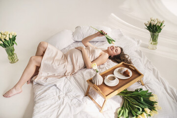 Fototapeta na wymiar An attractive pregnant woman in a silk peignoir on a bed among flowers, bouquets of tulips in a vase. Breakfast in bed. gentle studio pregnancy photo shoot