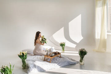 An attractive pregnant woman in a silk peignoir on a bed among flowers, bouquets of tulips in a vase. Breakfast in bed. gentle studio pregnancy photo shoot. Spring fresh