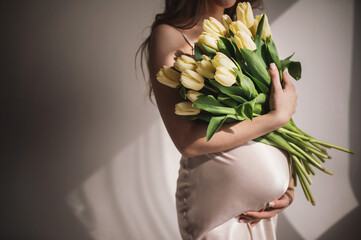 An attractive pregnant woman holds a bouquet of white tulips in her hands on a light background. Congratulations on Mother's Day, March 8th. Waiting for a miracle. tender pregnant curly shadows, hard 