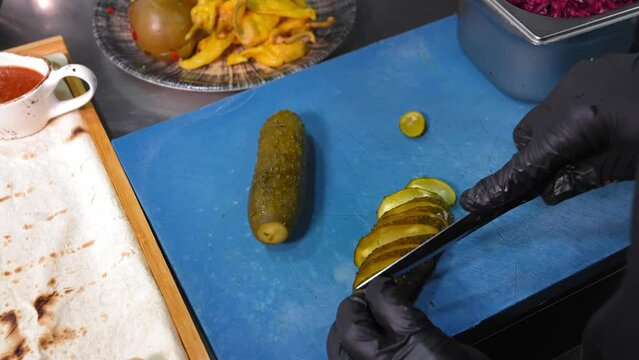 Chef prepares pickled cucumber salad. Pickled cucumber chef cuts with knife into pieces. Working environment on kitchen table next to container with pita bread and pickled vegetables, set.