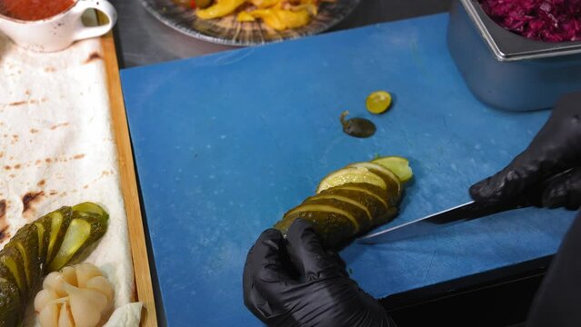 Chef prepares pickled cucumber salad. Pickled cucumber chef cuts with knife into pieces. Working environment on kitchen table next to container with pita bread and pickled vegetables, set.