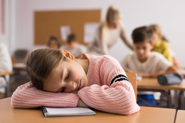 Portrait of tired school girl lying and sleeping at desk in classroom during lesson - obrazy, fototapety, plakaty