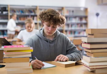 Intelligent male teenager engaged in research working with books in library