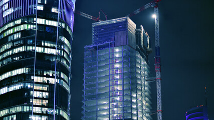Fototapeta na wymiar Skyscraper at construction process by night. Modern high-rise building under construction next to glass skyscrapers by night.