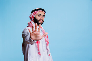 Muslim man wearing traditional clothes showing stop gesture with hand and looking at camera with...