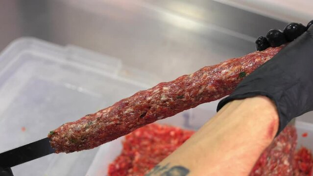 Chef sticks forcemeat on skewer and forming it for cooking lula kebab on grill at commercial kitchen. Close-up hands in black gloves