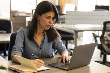 Businesswoman looking focused while taking notes and using a computer - obrazy, fototapety, plakaty