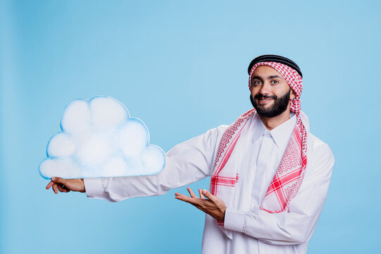 Smiling muslim man holding cloud shape and pointing with hand studio portrait. Person dressed in arab thobe and headscarf traditional clothes presenting weather forecast and looking at camera