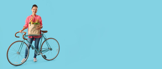 Young Asian woman with bicycle and bag full of fresh products on blue background with space for text