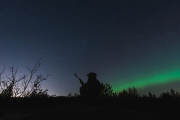 A special forces soldier with a night vision device and a rifle with a silencer is sitting in the forest against the background of the starry sky and northern lights.