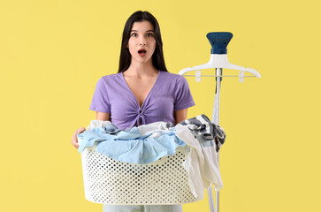 Shocked young woman with modern garment steamer and basket full of laundry on yellow background