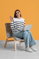 Beautiful young happy woman with laptop sitting on comfortable chair and waving hand near yellow wall