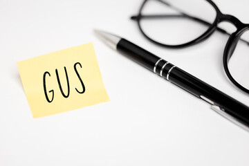 A yellow sticky note on a white background with the handwritten inscription "GUS", next to it a black pen and glasses (selective focus)