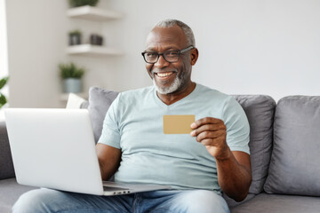 Smiling middle aged African gentleman sitting on the couch and using laptop and credit card to make online payment or transaction, online shopping or doing banking operation from home