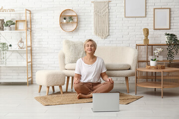 Mature woman with laptop meditating at home. Online yoga classes - Powered by Adobe
