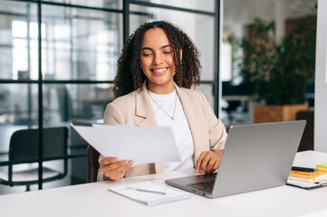 Positive clever curly brazilian or latino woman in elegant clothes, office employee, secretary or financial manager, sitting at her workplace with laptop in the modern office, studying documents smile