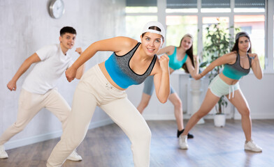 Fototapeta na wymiar Female teenager in ball cap engaged shake leg in fitness studio with friends, young people in good mood actively move and dance hip hop at choreography lesson