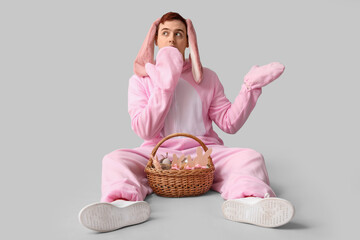 Surprised young man in Easter bunny costume sitting with basket on white background