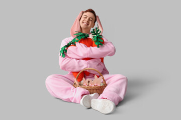 Happy young man in Easter bunny costume sitting with plush carrots and basket on white background