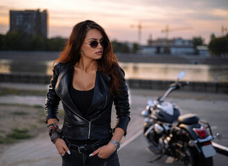 Attractive biker girl with sunglasses and leather jacket standing with her motorcycle in the background  © phoenix021