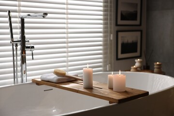 Wooden tray with burning candles, towel and brush on bathtub in bathroom