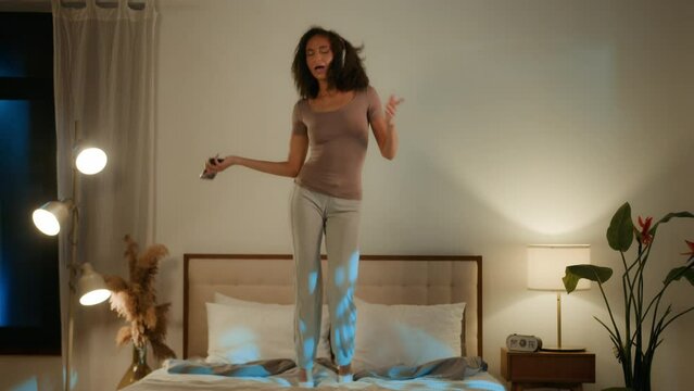 Happy African American teenager girl jumping on soft bed in bedroom active funny hilarious young ethnic woman listen music in headphones on phone singing dancing home party having fun in night weekend