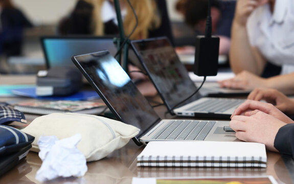 Busy school students working at a table on laptop computers with typical lesson accessories such as chargers, books, pens, rubbish and pencil cases.