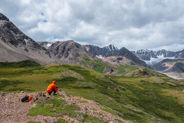 Photographer in vivid red wind jacket with hood and camera in hands in high mountains. Guy on stony hill takes pictures in scenic alpine valley with view to large glacier in sunlight under cloudy sky.