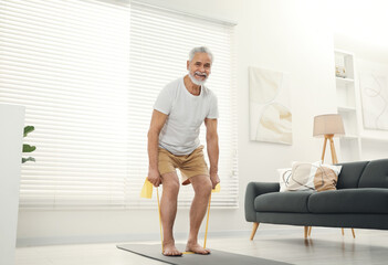 Senior man doing exercise with fitness elastic band on mat at home - 756100399