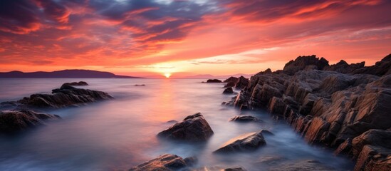 A picturesque natural landscape featuring a sunset over the ocean with rocks in the foreground, under a colorful sky with afterglow reflections on the water