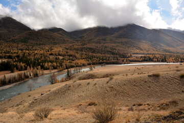 View from the top of the hill at the bend of a beautiful river flowing through the autumn valley at the foot of the mountains with peaks in the clouds.