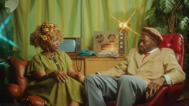 Cheerful African-American TV host smiling and talking with beautiful actress during interview on retro TV show in vintage studio