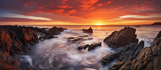 The afterglow of a stunning sunset paints the sky over a rocky shoreline, with waves crashing against the rocks in this picturesque coastal natural landscape