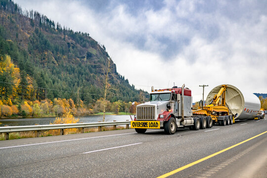 Big rig semi truck with oversize load sign on the bumper transporting oversized part of a wind turbine strut using an additional transport trolley running on the highway road along the Columbia River