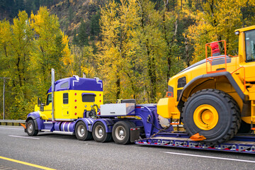 Blue and yellow powerful big rig semi truck transporting oversize load on heavy duty step deck semi trailer driving on the autumn highway road