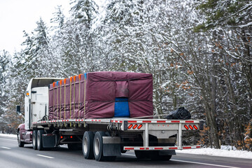 Classic big rig semi truck tractor transporting covered and fastened cargo on flat bed semi trailer...