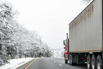 Red classic day cab big rig semi truck transporting cargo in long container driving on the slippery winter road with snowy forest on the sides
