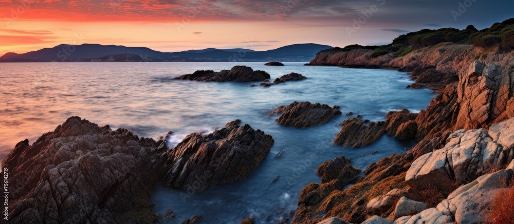 Wall mural A rocky cliff overlooking the fluid water of the ocean at dusk, with the sky painted in shades of pink and orange, creating a calming natural landscape