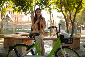 Naklejka premium Beautiful woman taking a bike stroll in the park, business woman holding smartphone using bike rental, business woman holding smartphone using bike rental digital phone