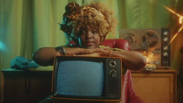 Young beautiful African-American woman resting head on old TV with static noise and posing at camera in studio with retro-styled interior. Video portrait, tracking shot