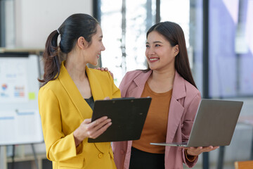Two pretty young Asian businesswomen standing to discuss working on investment projects and planning strategies together at the office. brainstorming concept.