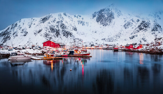 Red wooden houses against the background of a giant snow-capped peak. Majestic winter cityscape of Ballstad port, Norway. Spectacular spring view of Lofoten Islands. Calm seascape of Norwegian sea.