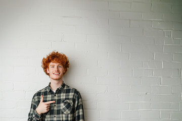 jeune homme souriant roux aux cheveux bouclés et portant une chemise bucheron verte, qui pointe du doigt vers l'espace négatif du fond, copyspace, fond mur briques blanches - obrazy, fototapety, plakaty