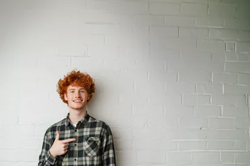 Foto op Plexiglas jeune homme souriant roux aux cheveux bouclés et portant une chemise bucheron verte, qui pointe du doigt vers l'espace négatif du fond, copyspace, fond mur briques blanches © Noble Nature
