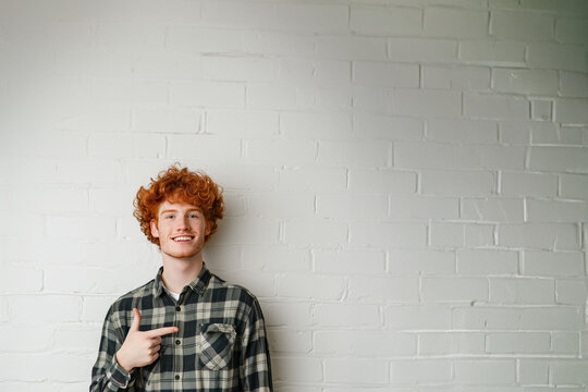 jeune homme souriant roux aux cheveux bouclés et portant une chemise bucheron verte, qui pointe du doigt vers l'espace négatif du fond, copyspace, fond mur briques blanches