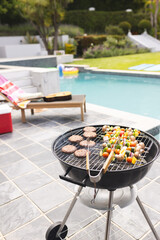 A charcoal grill is cooking burgers and vegetable skewers by a poolside with copy space