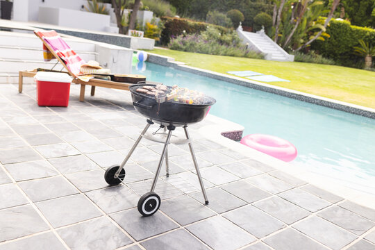 A charcoal grill is smoking beside a pool with a lounge chair and pink float