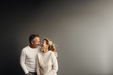 couple hétérosexuel quinqua, 50 ans environ, habillé avec des pulls blancs, qui se tiennent dans les bras et rigolent en se regardant. Fond gris avec espace négatif pour texte copyspace - obrazy, fototapety, plakaty