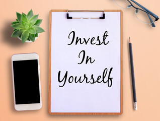 Top view text invest in yourself on clipboard, smartphone, pot plant and pencil on orange background.